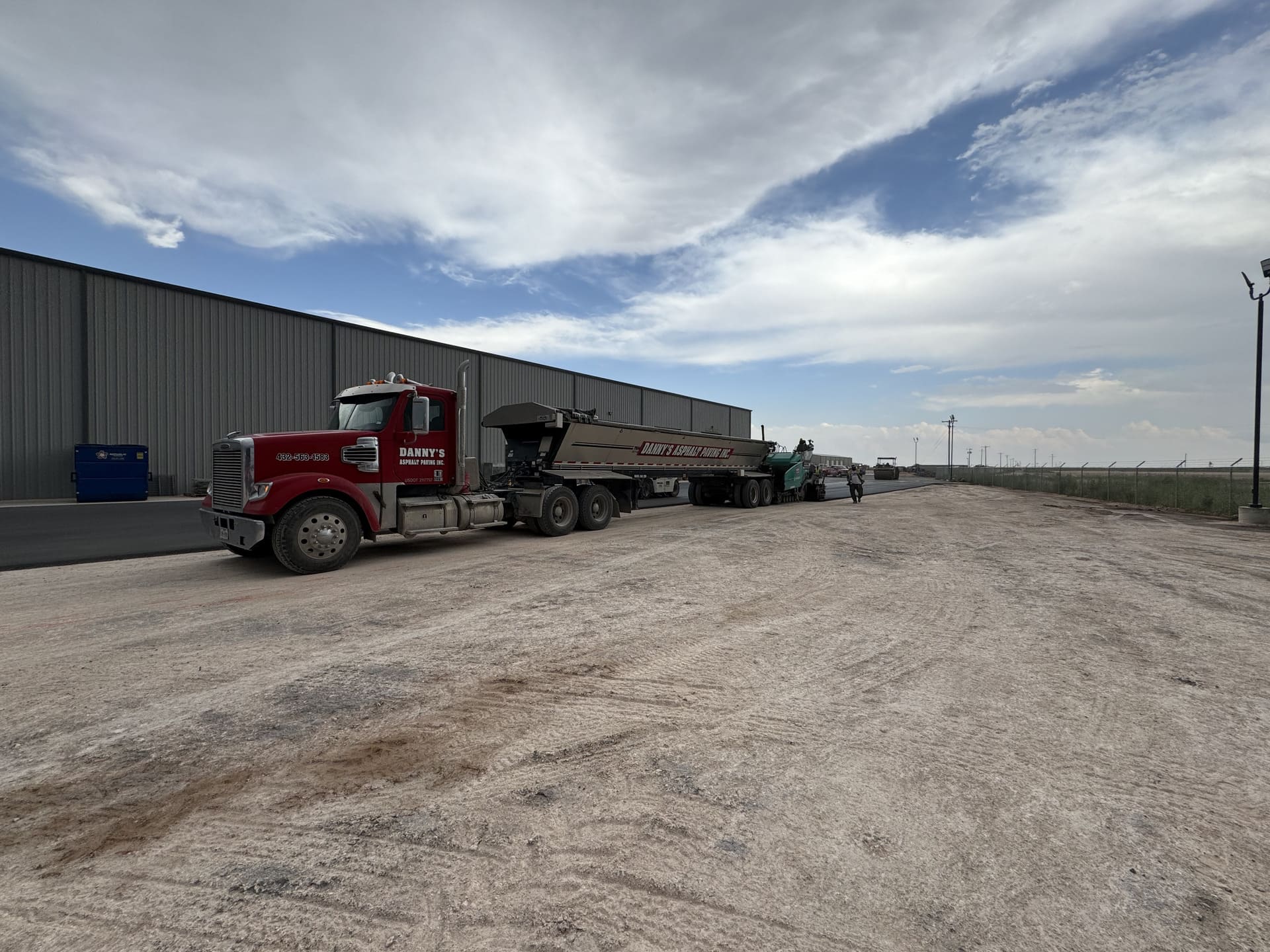 Danny’s Asphalt paving crew on-site in Andrews, TX with a red semi-truck and live bottom trailer preparing for industrial asphalt paving beside a large warehouse under a partly cloudy sky.