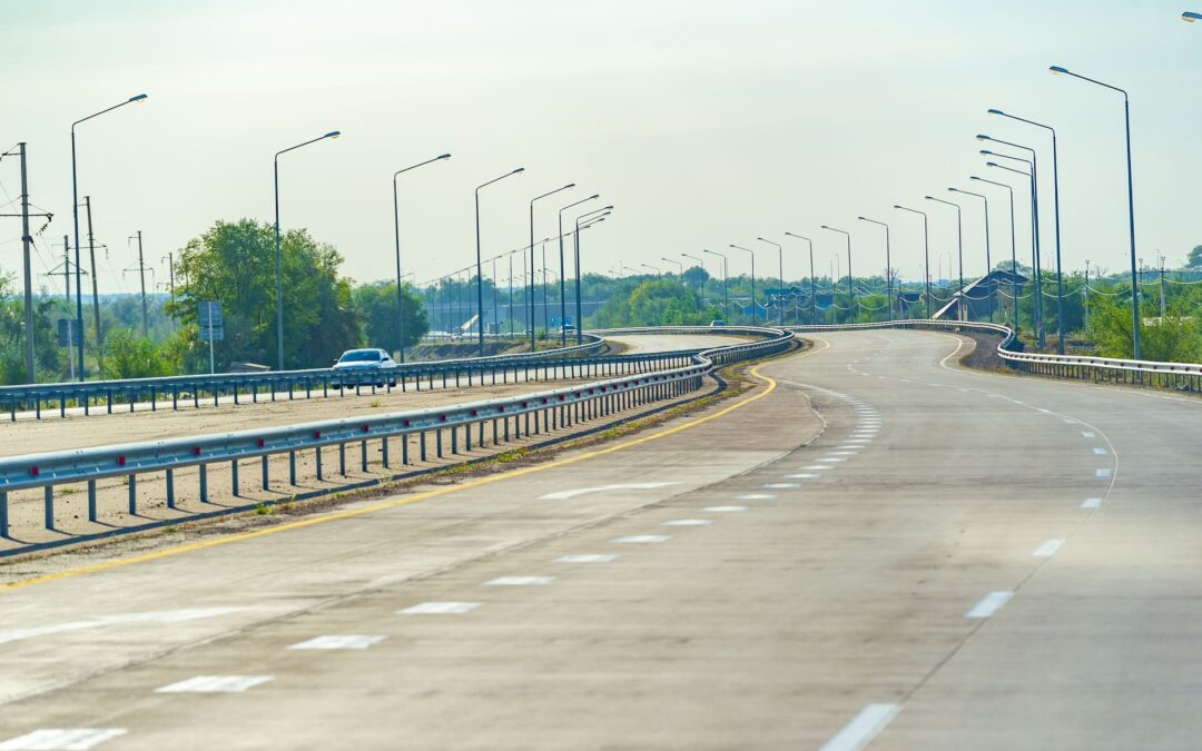 Wide-angle view of a freshly paved municipal asphalt road with clear lane markings, streetlights, and guardrails, set against a sunny sky and green roadside landscape.