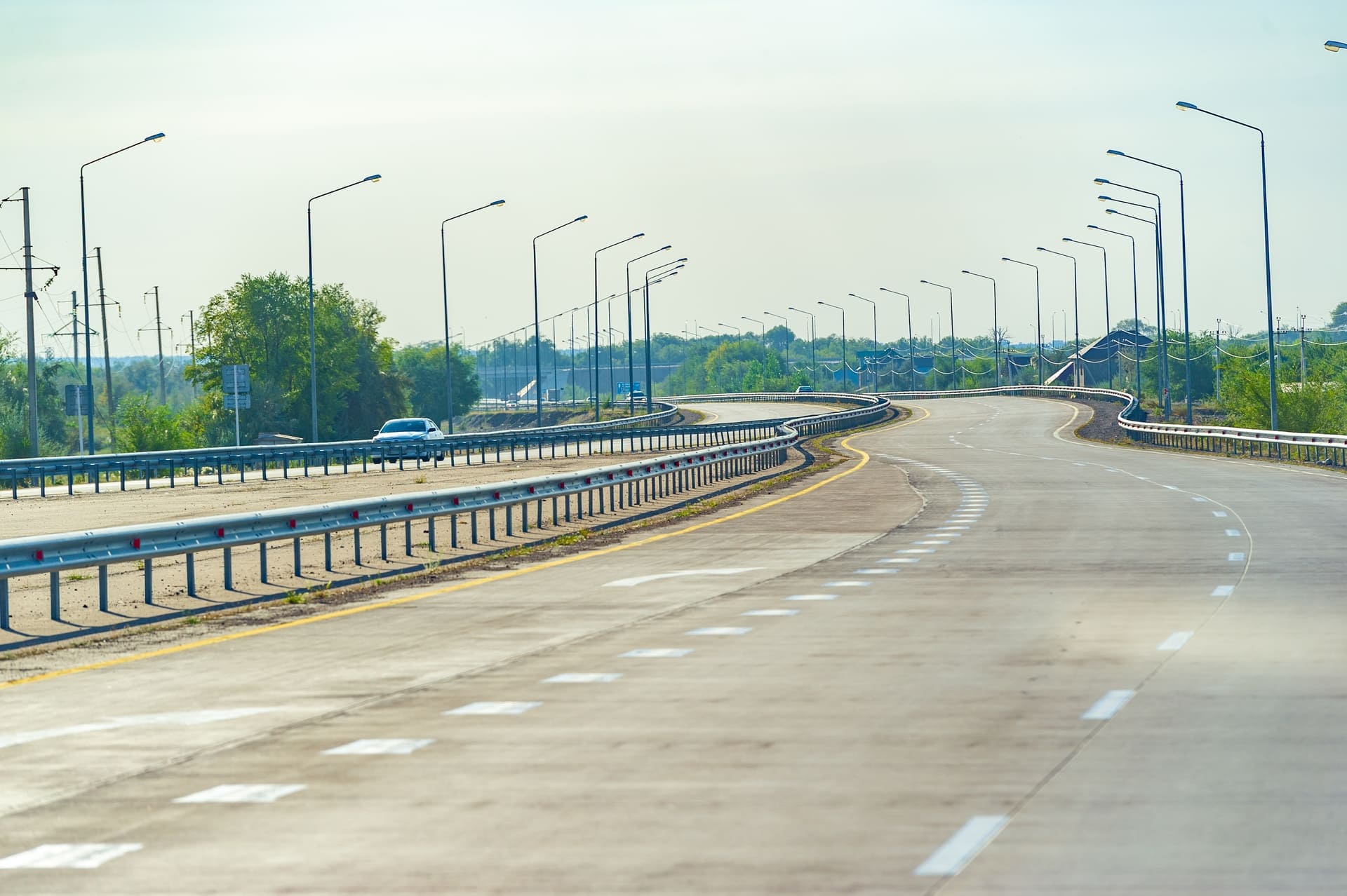 Wide-angle view of a freshly paved municipal asphalt road with clear lane markings, streetlights, and guardrails, set against a sunny sky and green roadside landscape.
