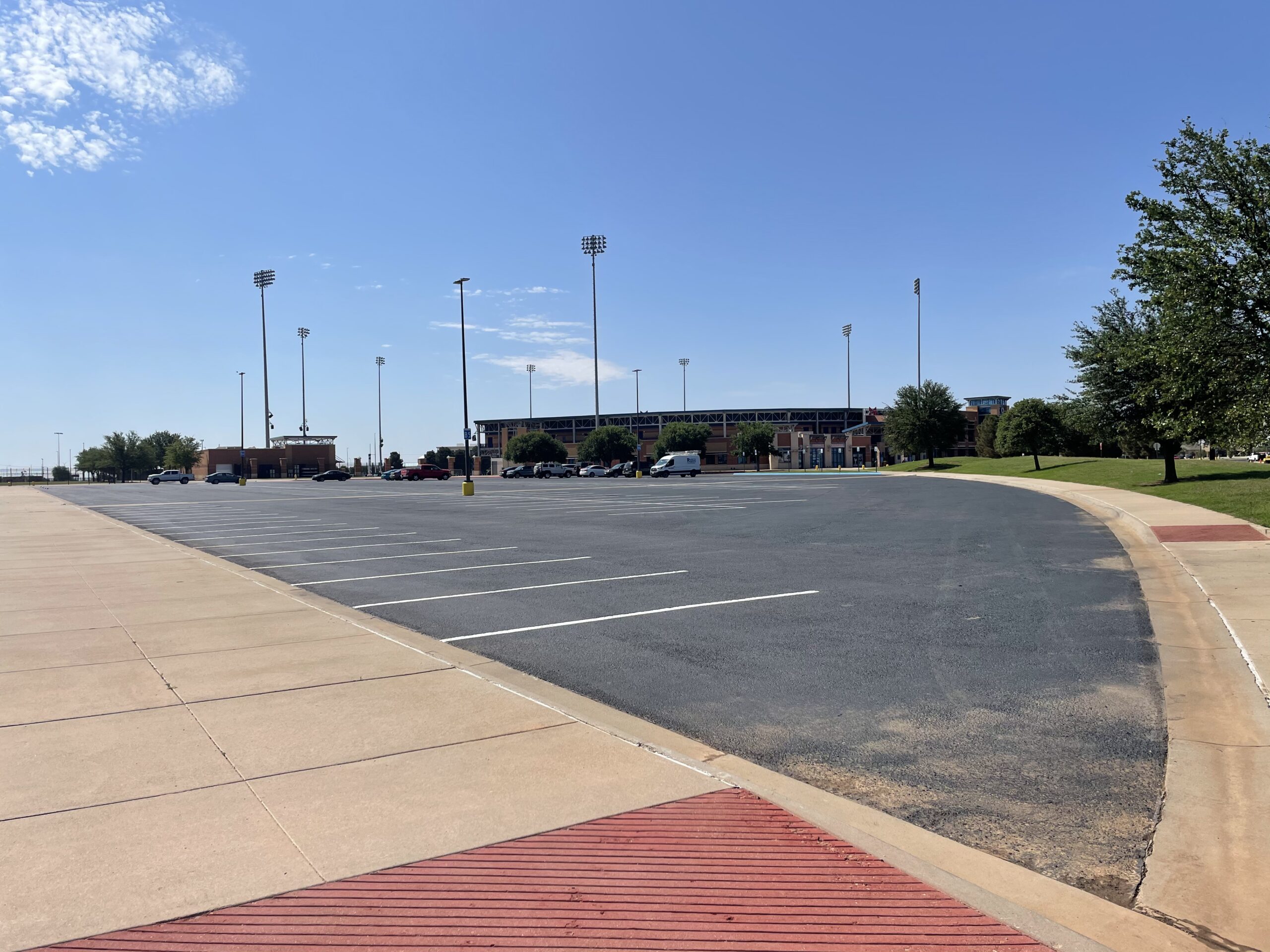 A wide-angle view of a freshly paved municipal asphalt parking lot in Andrews, TX, featuring crisp striping, smooth blacktop, and concrete sidewalks. Stadium lights and a public facility can be seen in the background under a clear blue sky, highlighting the scale and quality of the paving work.