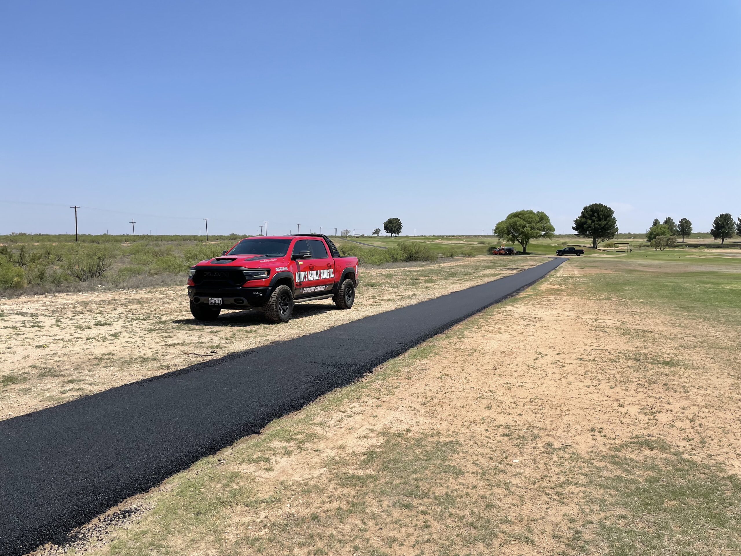 Red Danny’s Asphalt Paving Inc. pickup truck parked beside a freshly laid narrow strip of asphalt pathway in a rural area with dry grass, sparse trees, and a clear blue sky overhead.