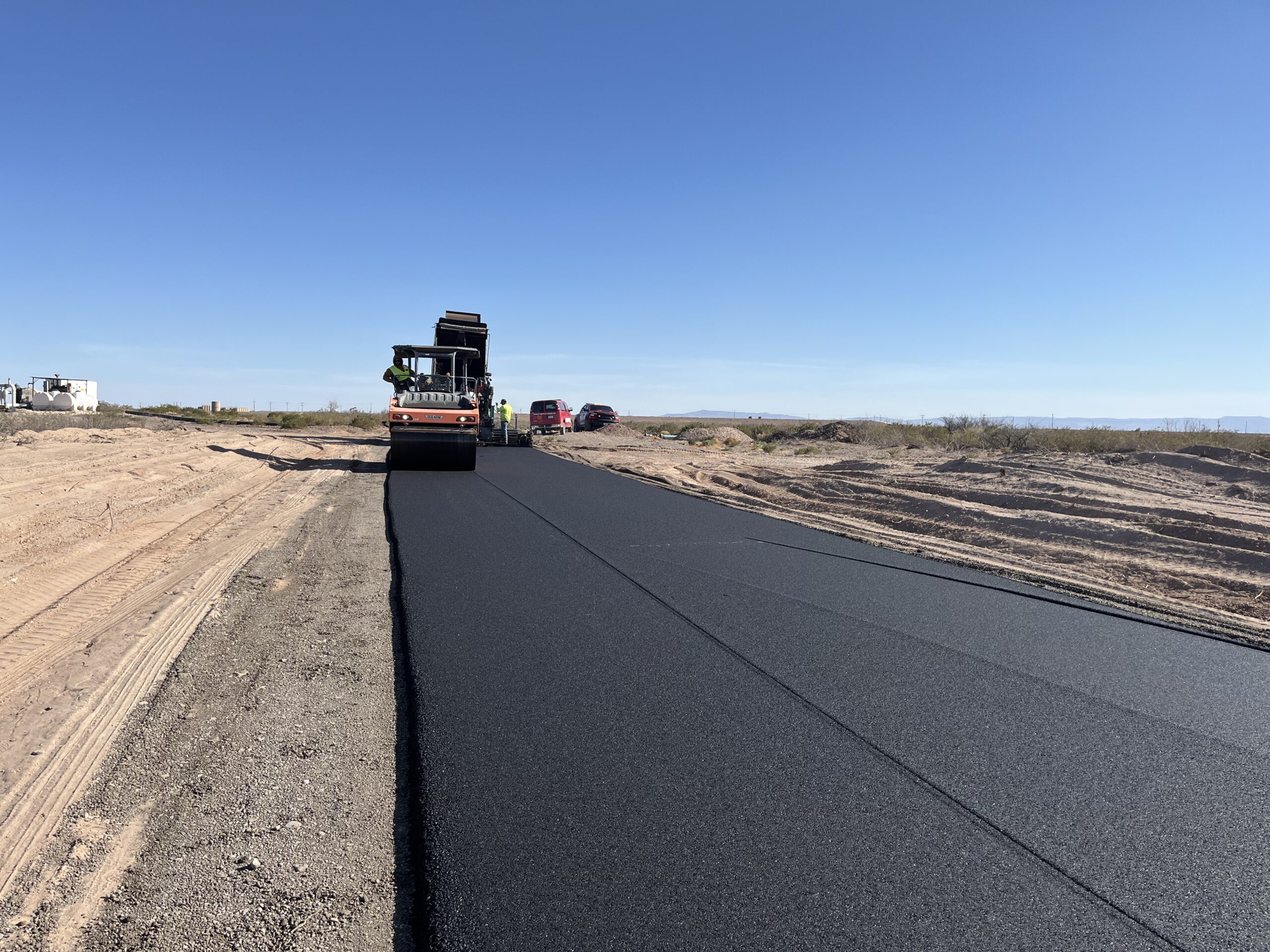 A freshly paved asphalt roadway stretches across a sandy construction area in Andrews, TX. A paving machine is laying the asphalt while workers and trucks follow behind under a clear blue sky, showcasing active municipal roadwork in progress.