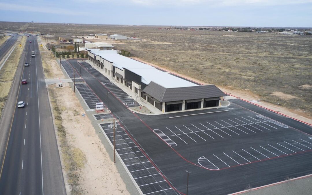 Aerial view of a freshly paved commercial parking lot adjacent to a modern retail strip in Midland/Odessa, Texas. The asphalt is newly striped with white and red markings, and the surrounding area shows wide-open West Texas terrain and a nearby highway with light traffic