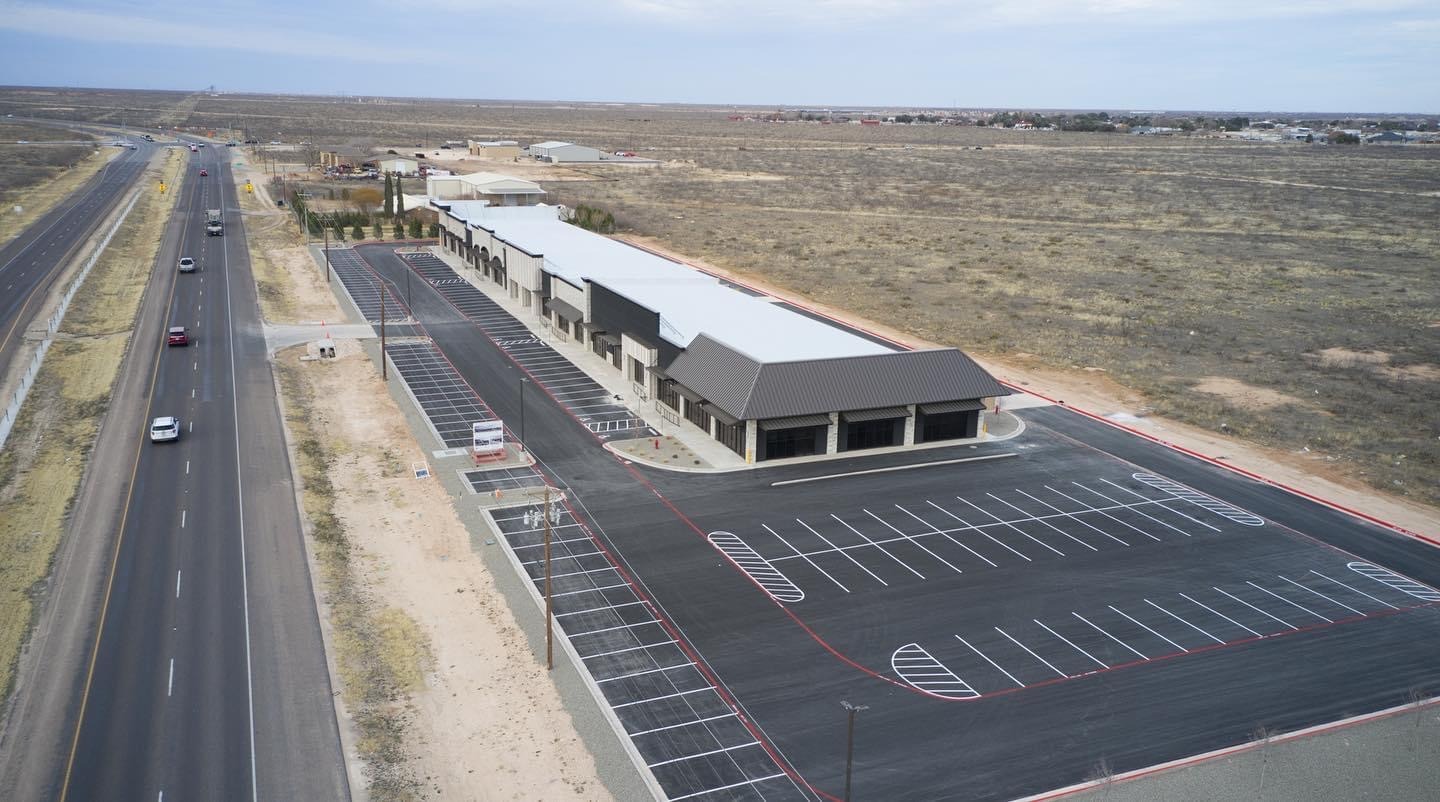 Aerial view of a freshly paved commercial parking lot adjacent to a modern retail strip in Midland/Odessa, Texas. The asphalt is newly striped with white and red markings, and the surrounding area shows wide-open West Texas terrain and a nearby highway with light traffic