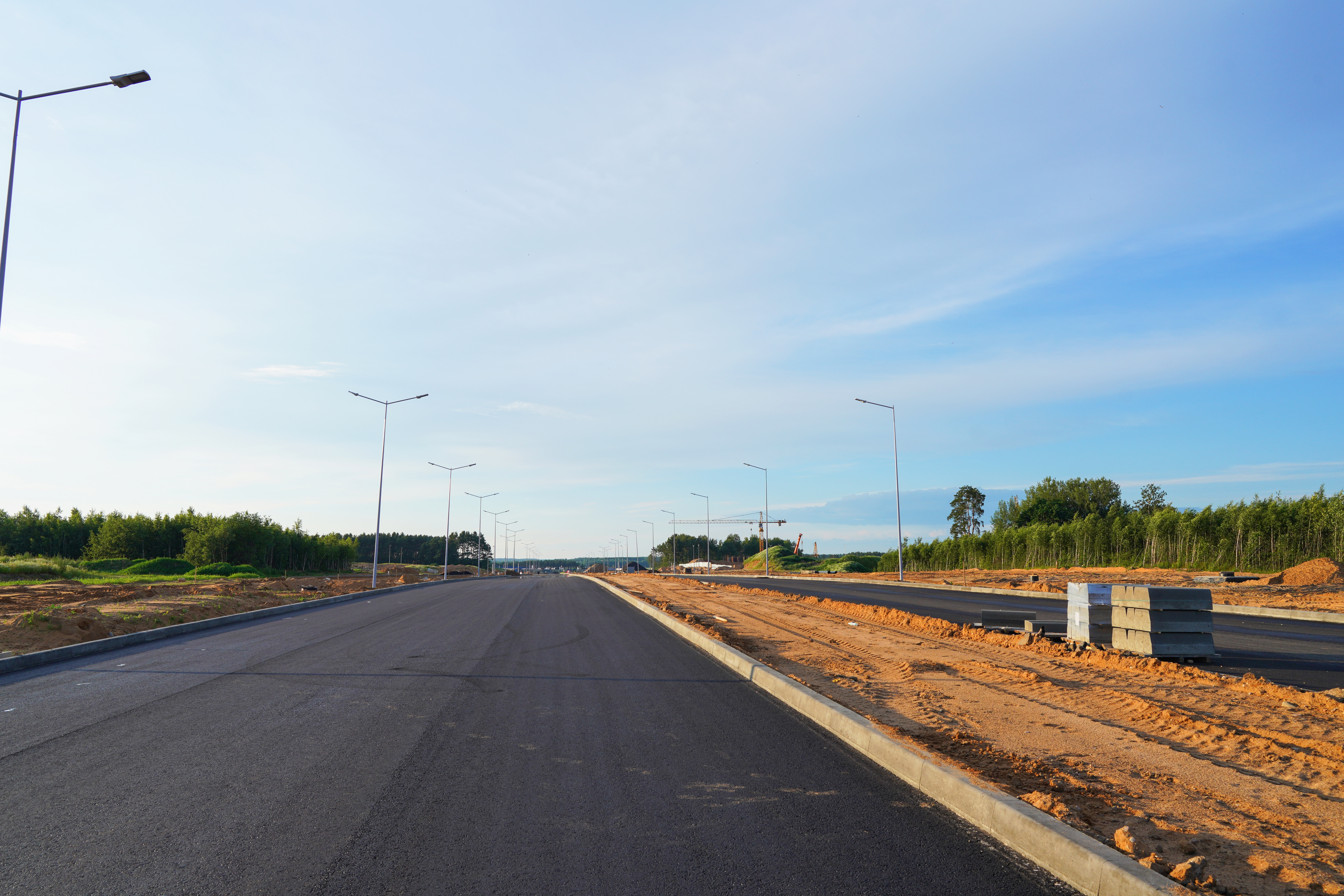 Freshly paved asphalt road under construction with new light poles installed, bordered by dirt and greenery, under a clear blue sky in Andrews, Texas.