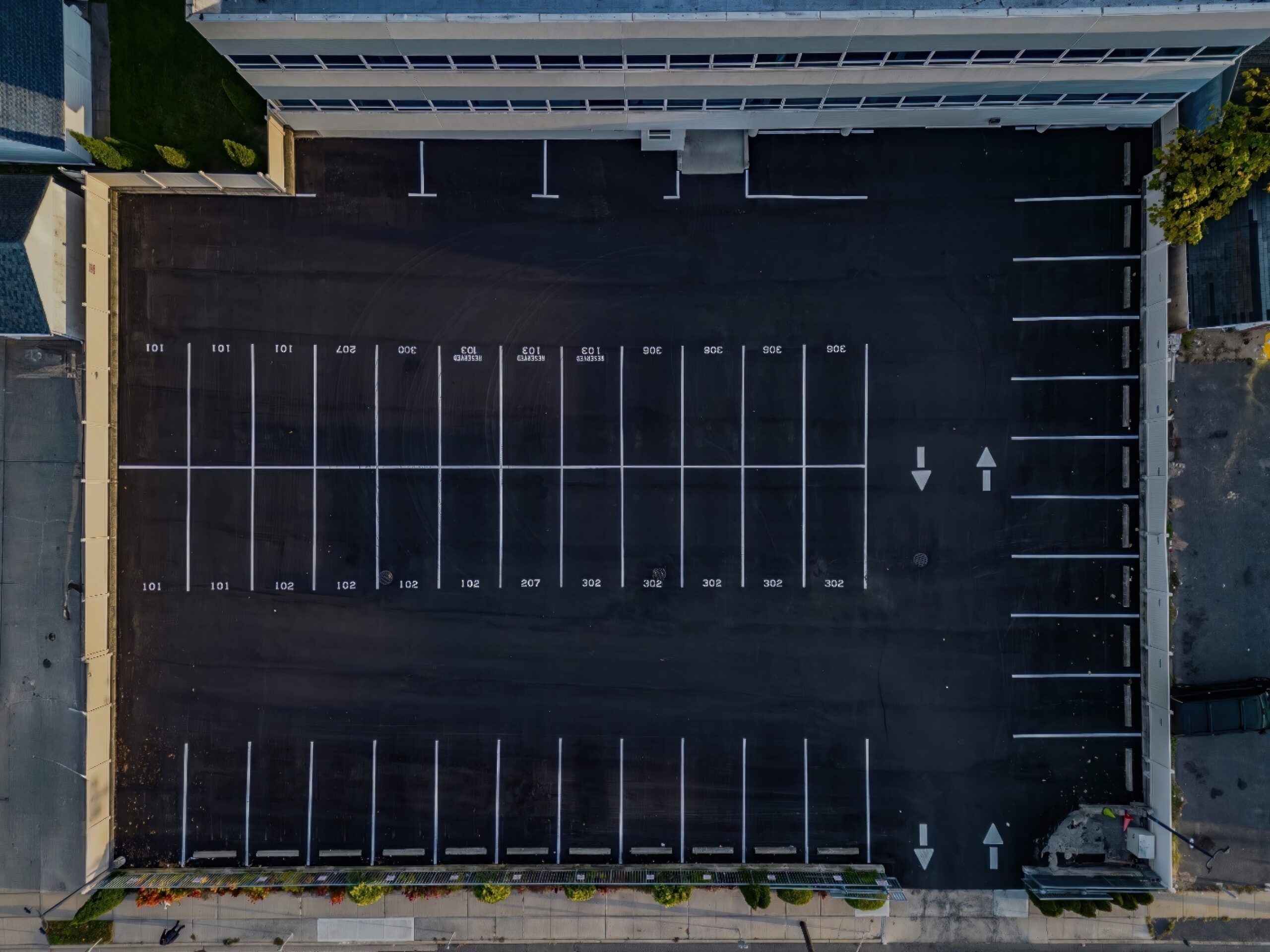 Aerial view of a freshly paved institutional asphalt parking lot with clearly marked white parking lines, directional arrows, and designated parking stalls adjacent to a commercial or educational building.