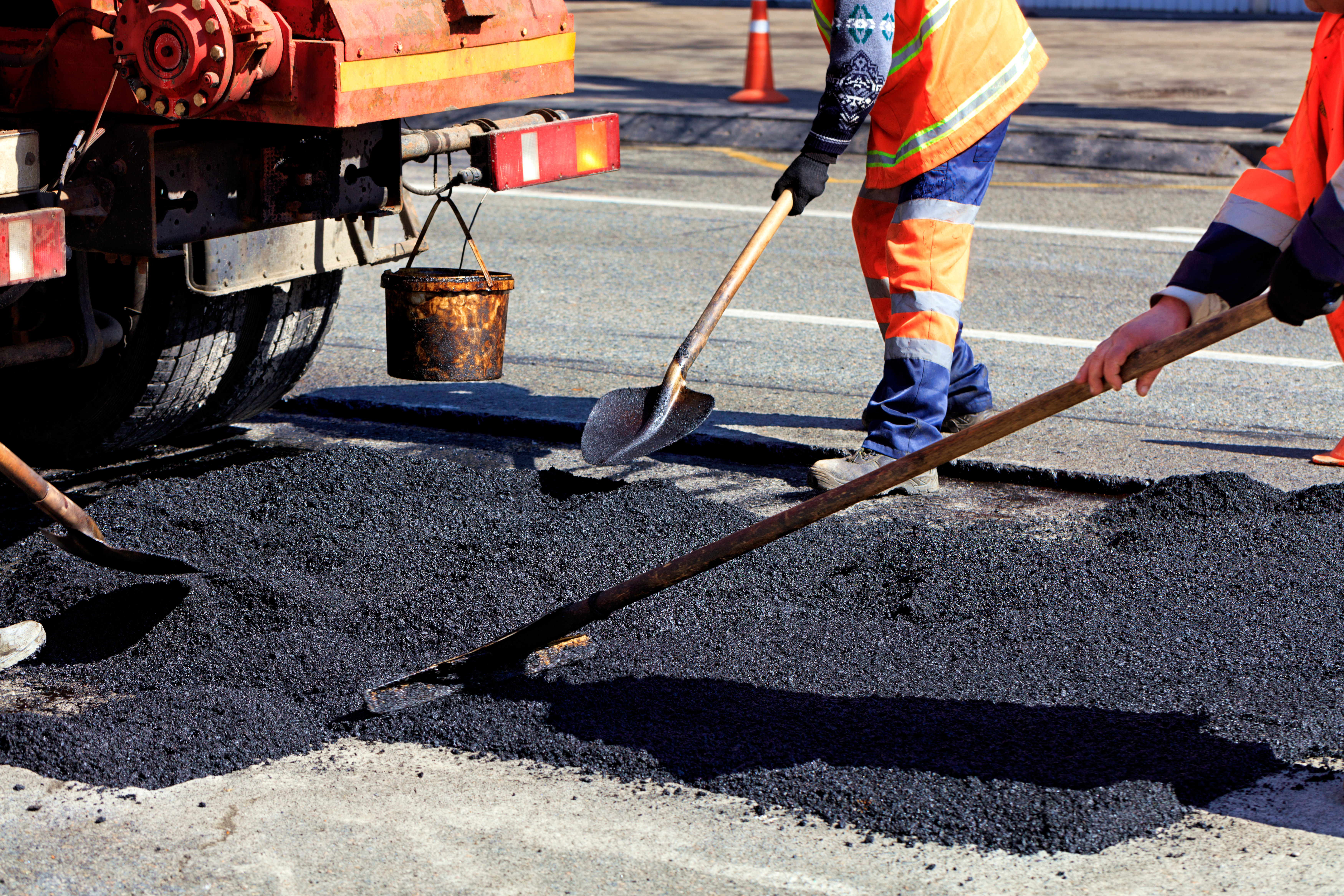 Workers in high-visibility gear performing asphalt patch repair on a road using shovels and rakes beside a paving truck during daylight.