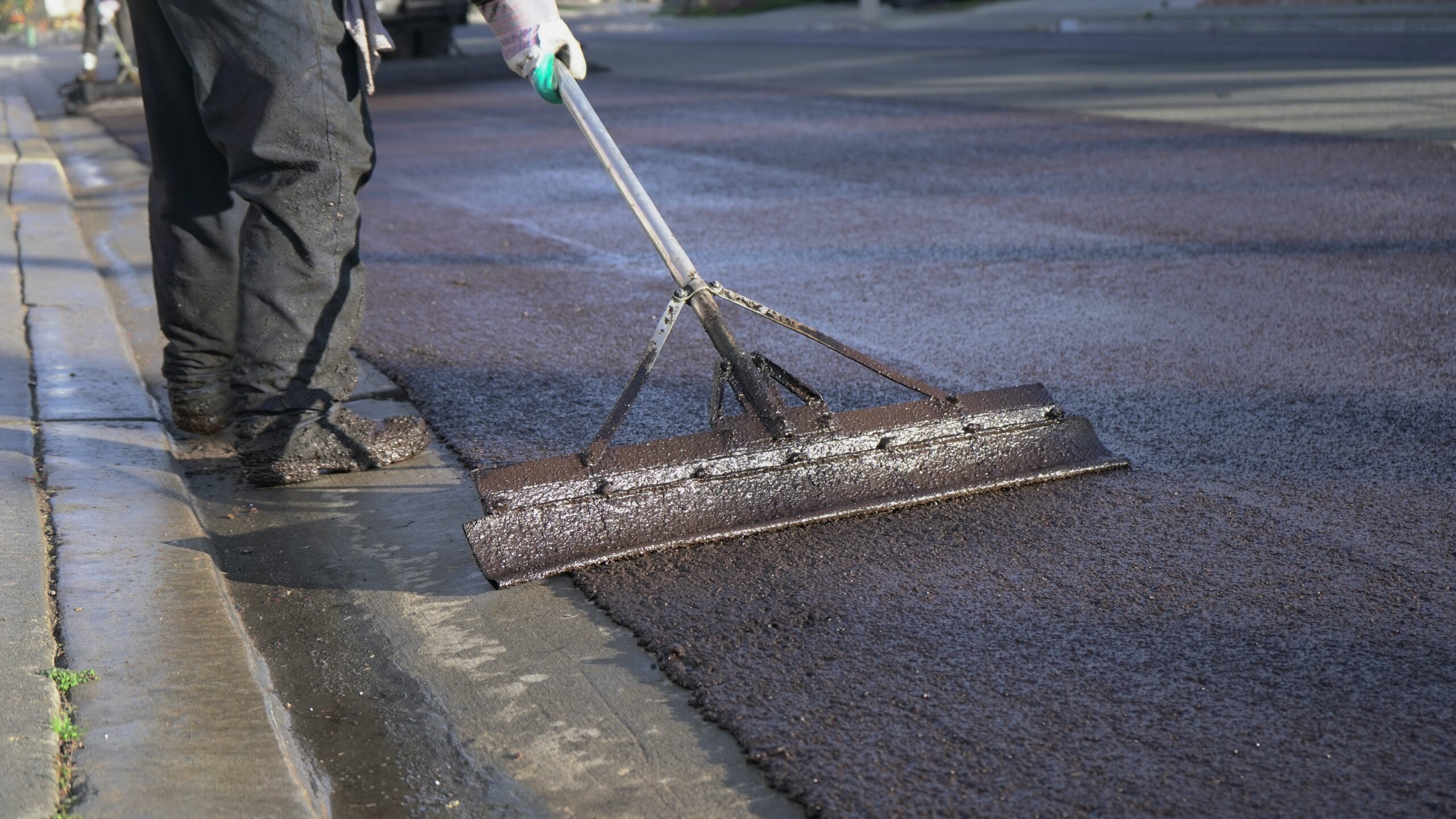 Worker applying sealcoat to a paved asphalt road using a large squeegee tool along the curb edge, highlighting pavement maintenance in progress.