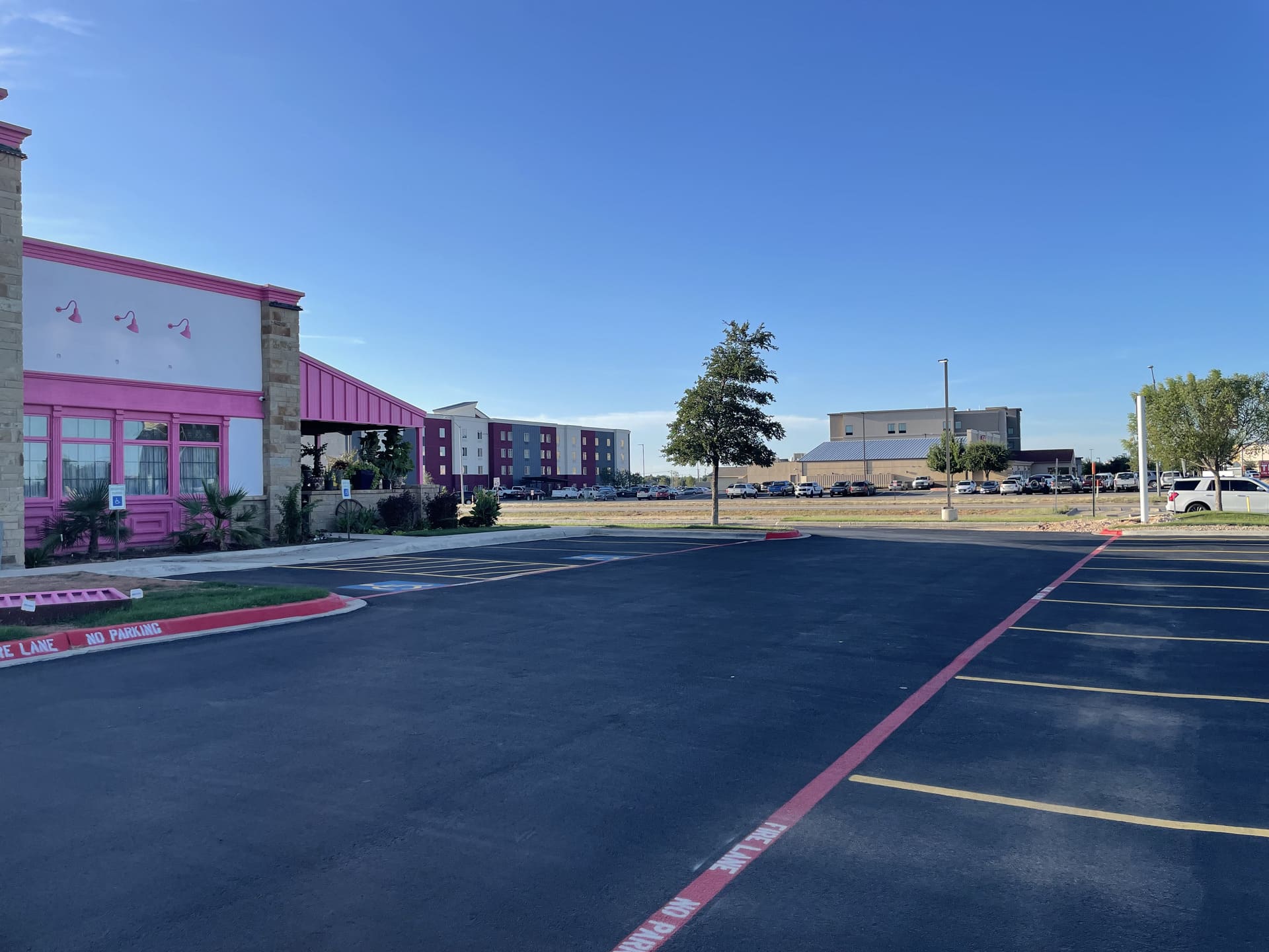 Professional Asphalt Paving Freshly paved commercial parking lot in Andrews, Texas, featuring smooth black asphalt, bright yellow parking lines, and red fire lane markings beside a colorful storefront under a clear blue sky.