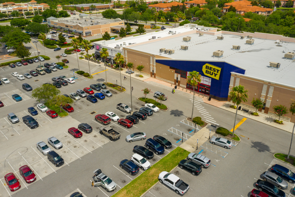 Aerial view of a freshly paved institutional asphalt parking lot with clearly marked white parking lines, directional arrows, and designated parking stalls adjacent to a commercial or educational building.