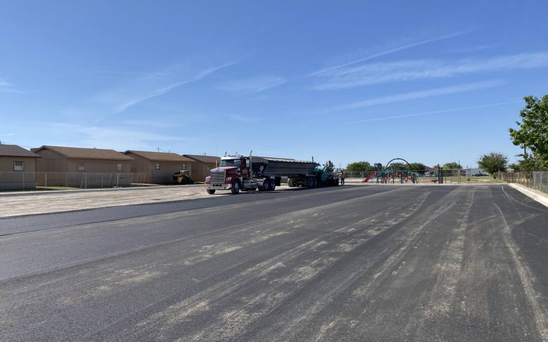 New asphalt being laid in a residential subdivision, with paving equipment and a dump truck working near homes and a neighborhood playground.