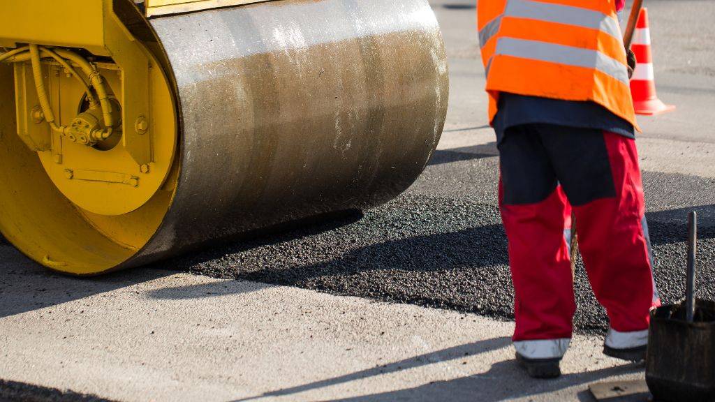 Heavy steel drum roller compacting hot-mix asphalt during an industrial base repair project.