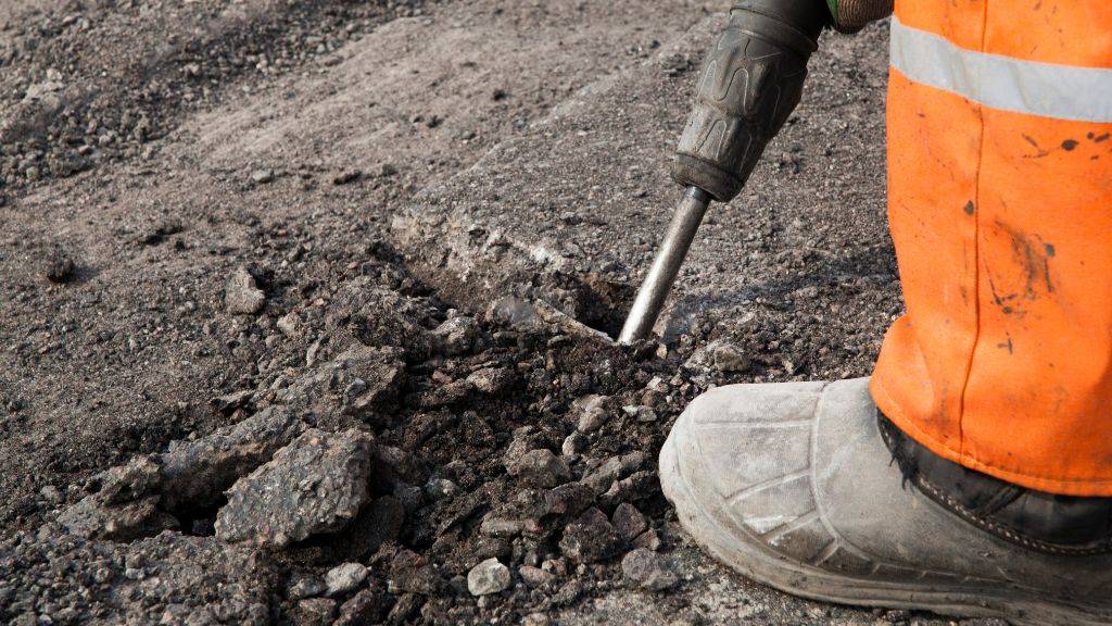 Construction worker using a jackhammer to remove damaged pavement for industrial base repairs before winter freeze-thaw cycles.