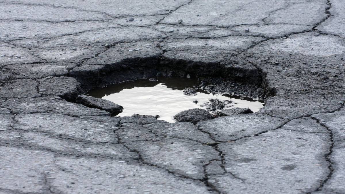 Large pothole filled with water in cracked asphalt pavement caused by heavy traffic wear at a venue parking lot.